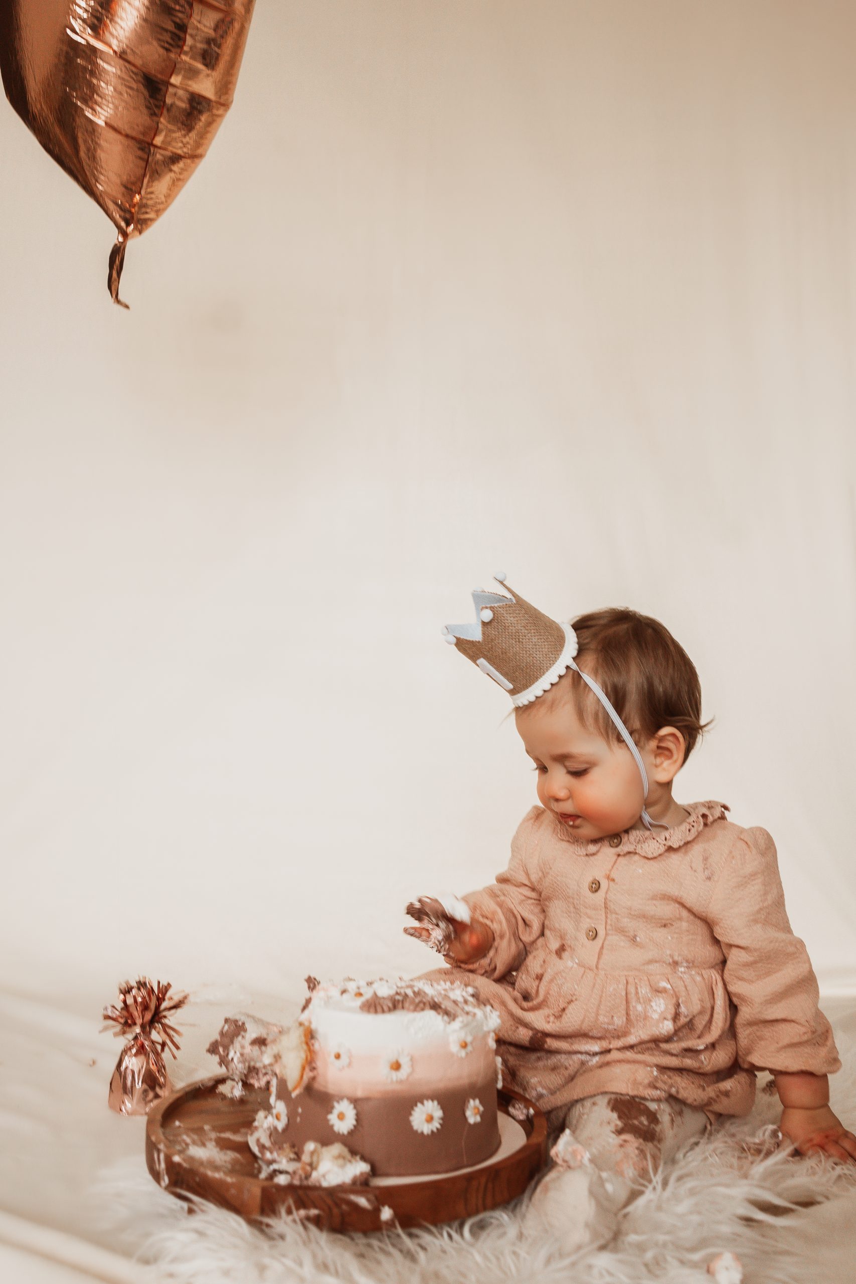 "Een liefdevol moment tijdens een eerste verjaardagsshoot, vastgelegd in warme, natuurlijke tinten. De baby draagt een oudroze jurkje en een handgemaakt kroontje, zittend op een zacht vloerkleed met een rustieke taart vol fijne details zoals margrietjes. De zachte styling met neutrale kleuren, ballonnen in goud en taupe en de dromerige sfeer weerspiegelen de pure, tijdloze stijl van ByIrisvanderZwan fotografie."