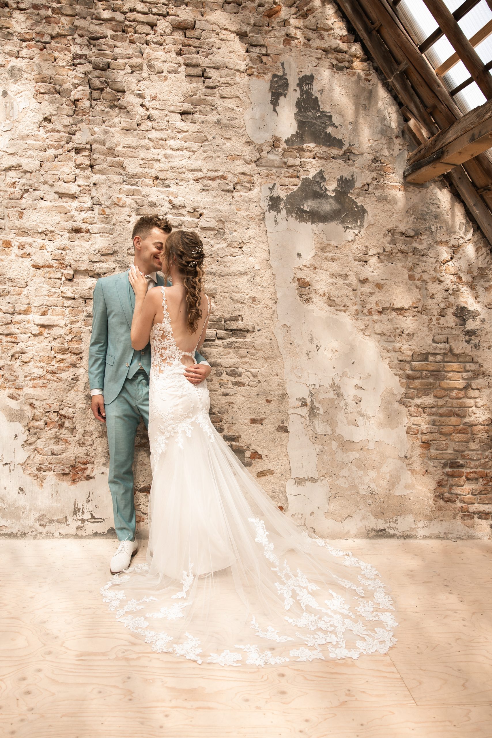 Romantisch moment tussen een bruid en bruidegom bij een groot raam, waarbij de bruidegom haar liefdevol een kus op het voorhoofd geeft. De bruid draagt een kanten off-shoulder jurk en houdt een boeket bloemen vast.