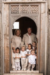 Grootouderfoto met vijf jonge kleinkinderen in zachte, neutrale tinten voor een houten sierdeur tijdens een warme familiefotoshoot in het Westland, ’s-Gravenzande, regio Den Haag, vastgelegd door ByIrisvdZwan