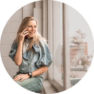 Professionele portretfoto van een lachende vrouw die telefoneert bij een groot raam, zachte beige en groene tinten, natuurlijk licht; moderne lifestylefotografie voor moeders en vrouwen in het Westland, ’s-Gravenzande en regio Den Haag door By Iris van der Zwan.