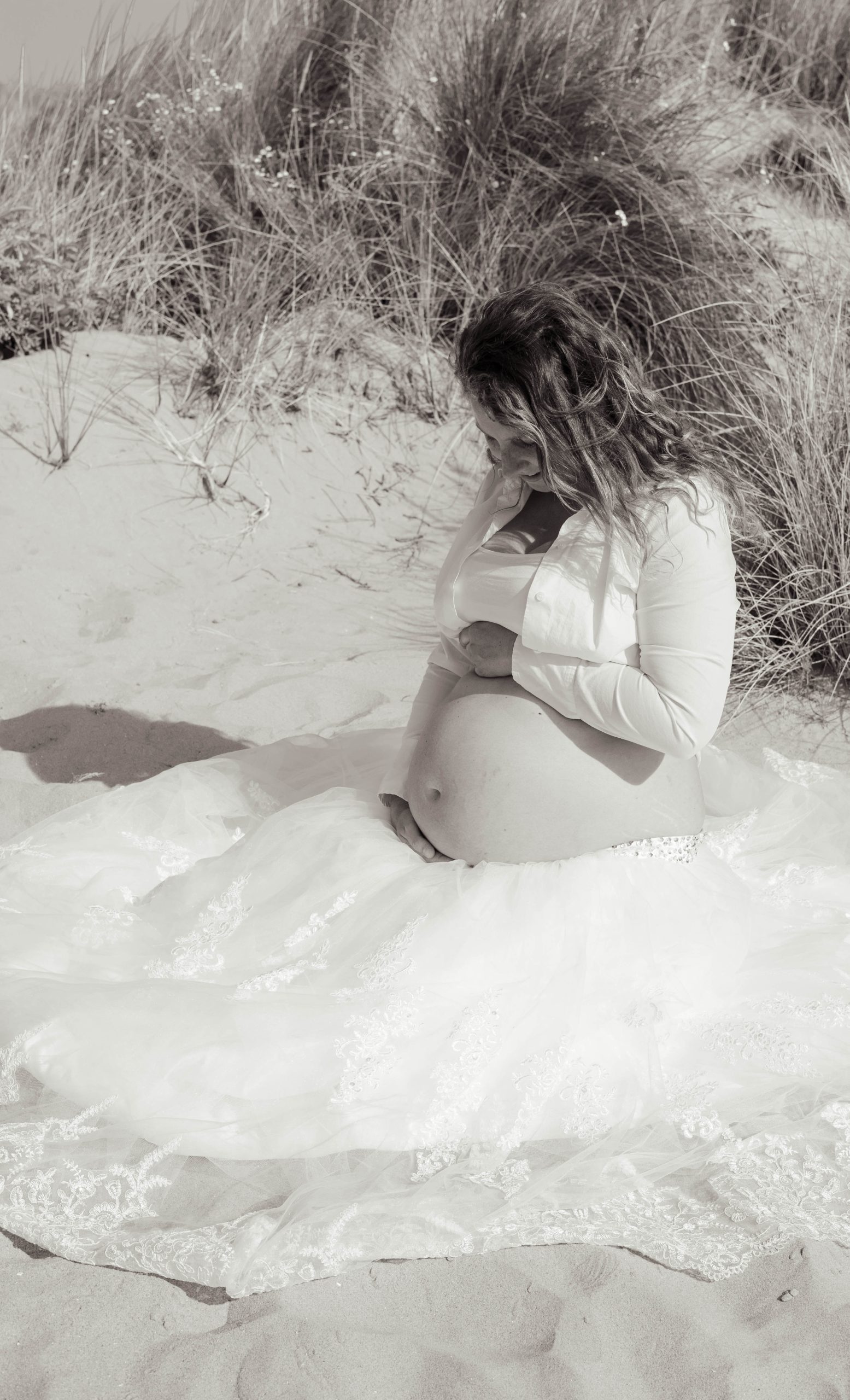 Zwangere vrouw zittend in het zand tijdens zwangerschapsshoot op het strand in het Westland, ’s-Gravenzande, regio Den Haag, door ByIrisvdZwan